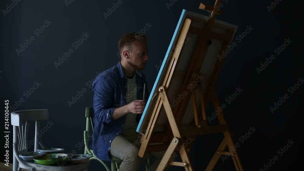 young male blond artist paints with a brush and oil paints in a dark workshop. The canvas young male blond artist paints with a brush and oil paints in a dark workshop. The canvas