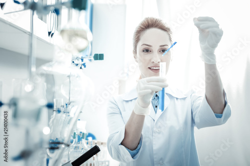 Professional biological research. Cheerful positive female biologist holding a test tube and looking at it while working on her project in the lab