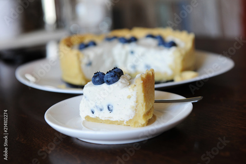 Piece of cheesecake with blueberries on a plate in focus on the background of whole cheesecake