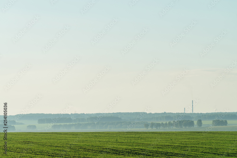 Misty fragment of plowed field in springtime. Rich green background of field with furrows from plough under blue sky with copy space. Tree in haze and industrial pipes with smog on horizon.