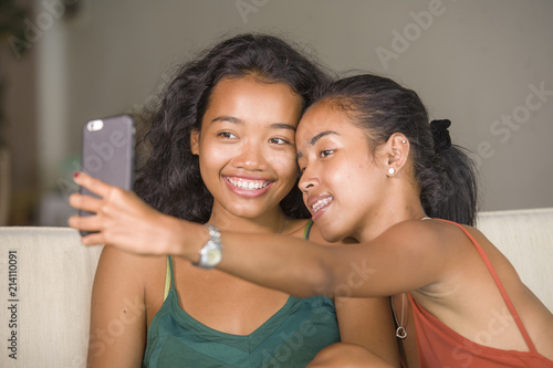 young happy and beautiful Asian sisters or girlfriends couple smiling cheerful taking selfie photo with mobile phone at home couch playful laughing together photo