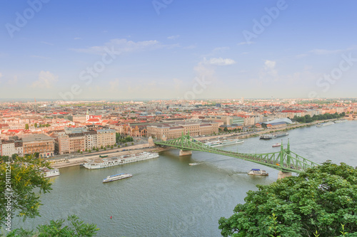 Budapest skyline with red roofs of the buildings and the Liberty Bridge across the Danube and boats sailing on the river.