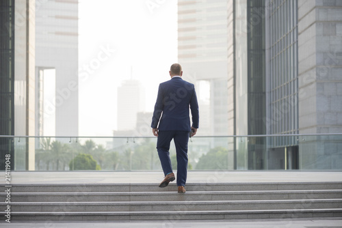 Rear view of white middle age, bearded corporate businessman wearing a suit  walking, up steps in Dubai Business district while holding his phone © Paul