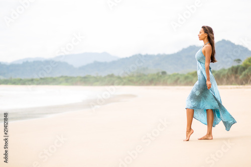 Fashion outdoor photo of gorgeous young sexy lady in long blue dress walking barefoot on the sea shore. Summer sunny day and holiday concept  seascape with girl  beach  beautiful waves  blue water.