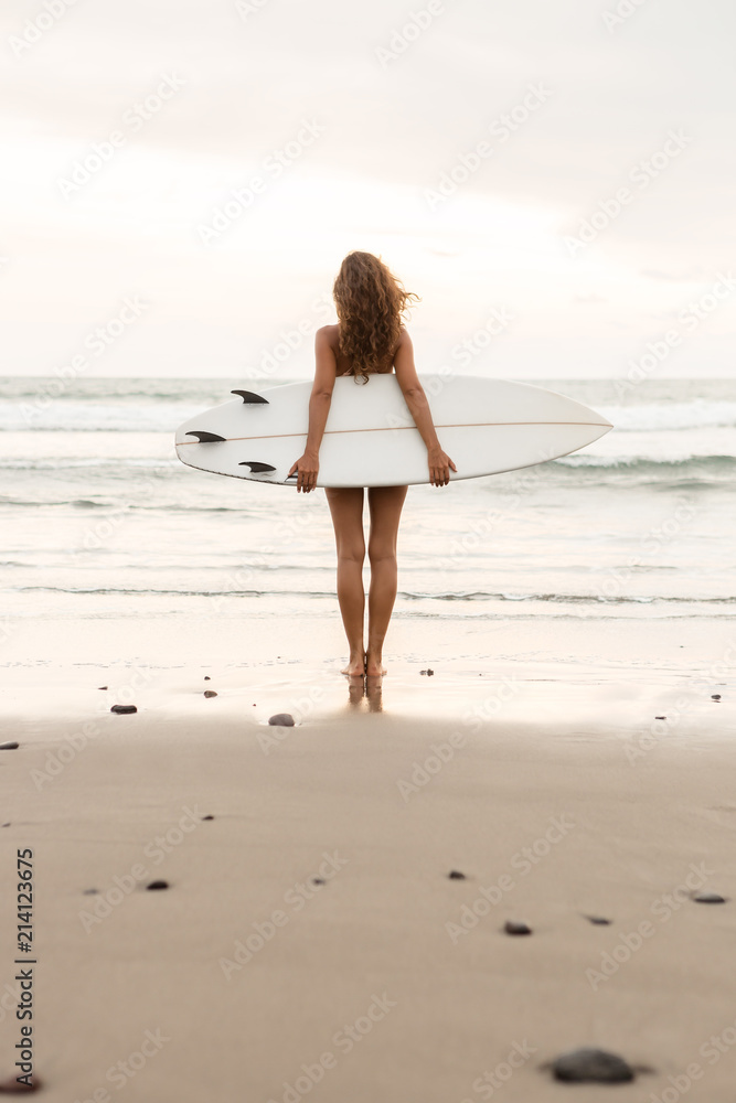 Surf girl with long hair go to surfing. Young surfer woman holding blank white short surfboard on a beach at sunset or sunrise. Bali island, Indonesia. Outdoor Active Lifestyle. It's time for surfing!