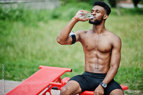 African american male athlete sexy sport bare torso man with running sports arm case for mobile phone  doing exercises on the street workout simulators and drinking water from bottle.