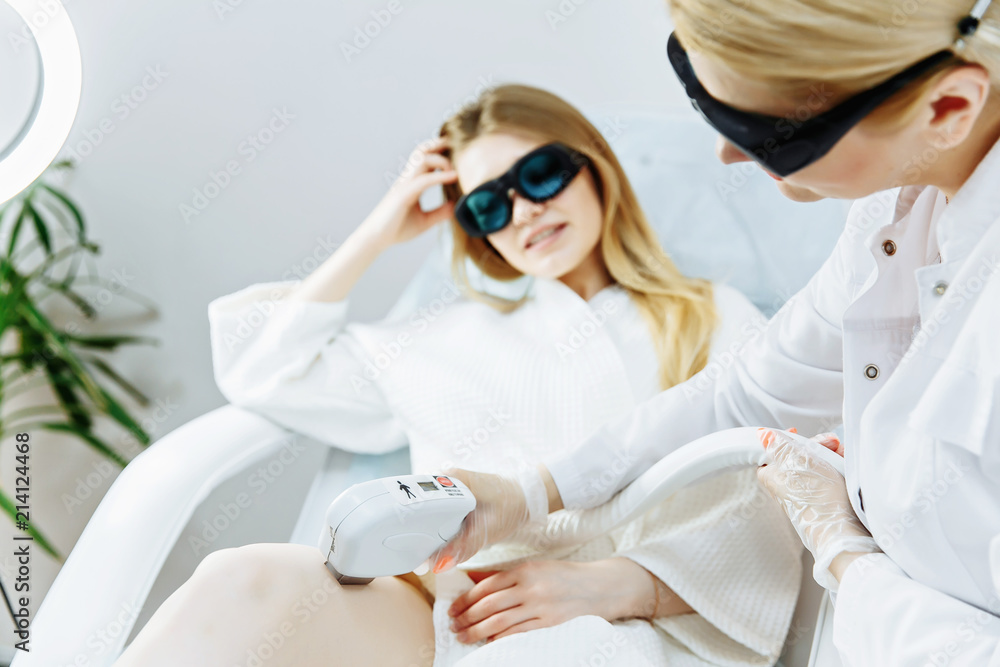 The girl is lying on the couch in the medical glasses in the treatment room. The hands of the cosmetologist make the epilation of the legs The girl is lying on the couch in the medical glasses in the treatment room. The hands of the cosmetologist make the epilation of the legs