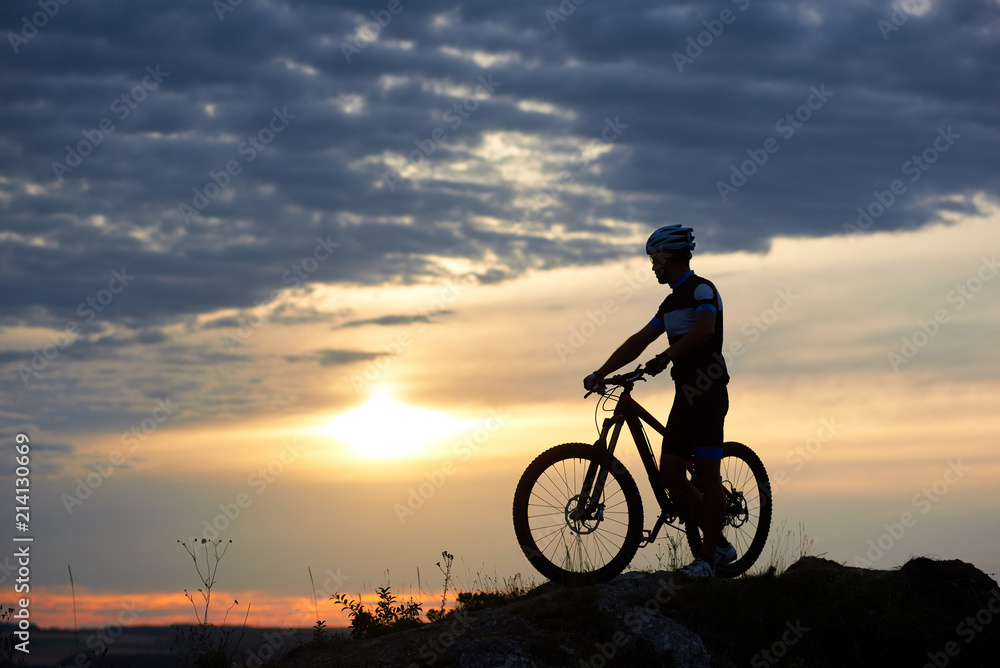 Fototapeta premium Athletic man in sportswear and helmet holding hands on bicycle and standing near it. Energetic cyclist posing in twilight and sunset on hill and against wonderful background of sky and clouds.