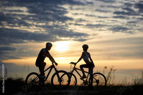 Man and woman cyclists on bicycles standing on the road among the field grass opposite each other and looking to the side at sunset. A magical sky with clouds among which the sun that sits is seen