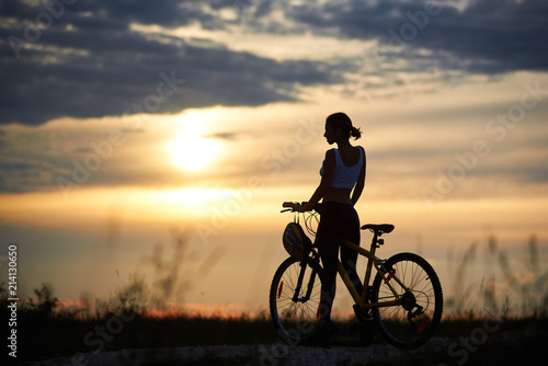 Silhouette of sporty cyclist  wearing sportswear and posing near her bicycle on trail. Slender incognito woman enjoying nature and observing wonderful landscapes and amazing sunset.