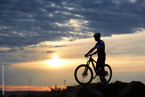Athletic man in sportswear and helmet holding hands on bicycle and standing near it. Energetic cyclist posing in twilight and sunset on hill and against wonderful background of sky and clouds.