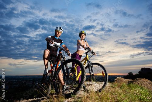 Young people on mountain bikes on top of a hill under a magic sky at sunset. The guys are dressed in sports clothes and helmets and look at the camera
