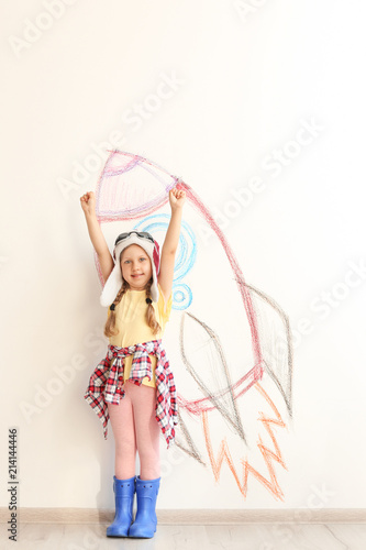 Adorable little child playing astronaut near wall with drawing of spaceship indoors