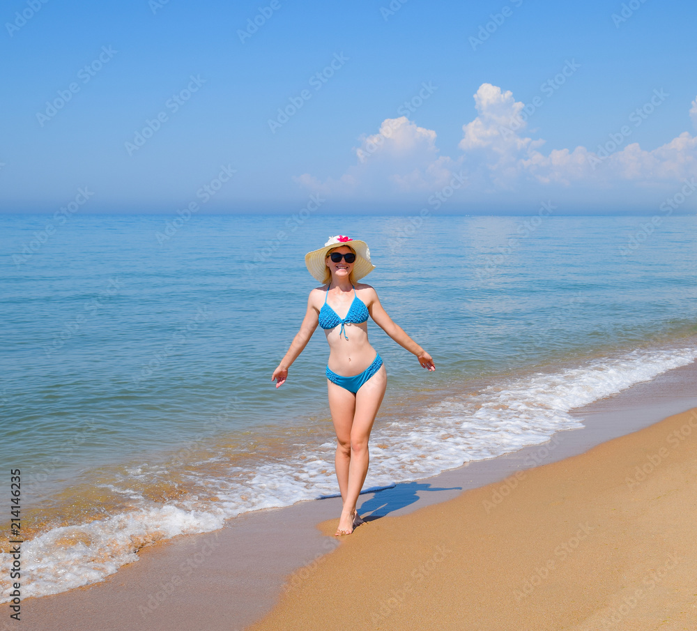 Blonde in a blue swimsuit and a white hat on the beach. Beautiful sea beach.