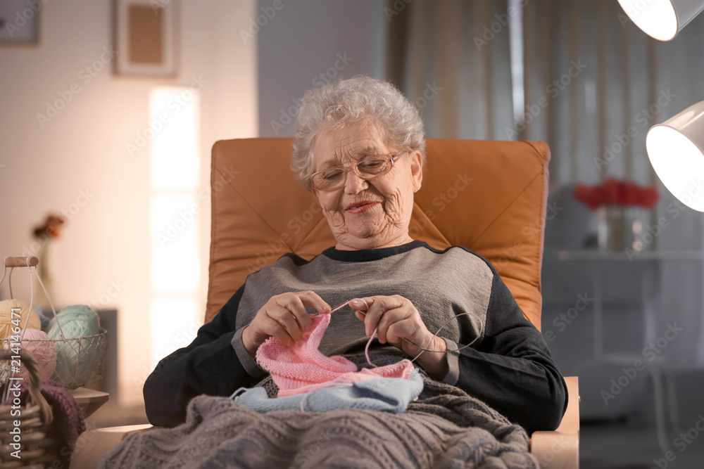 Senior woman knitting warm sweater in armchair at home - obrazy, fototapety, plakaty 