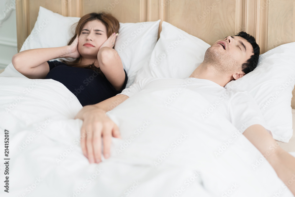 Annoyed Asian wife blocking her ears from noise of husband snoring in bedroom at home. Young couple have problem with man's snoring. Annoyed Asian wife blocking her ears from noise of husband snoring in bedroom at home. Young couple have problem with man's snoring.