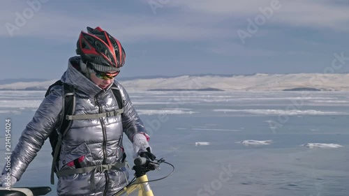 Woman is wearing sports equipment. The girl is dressed in a silvery down jacket, cycling backpack and helmet. Ice of the frozen Lake Baikal. The tires on the bicycle are covered with special spikes photo