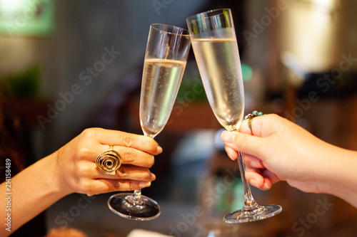 Cheers and Celebration. Two women hands with rings holding glasses of champagne making a toast with indoor background. Selective focus on left hand.