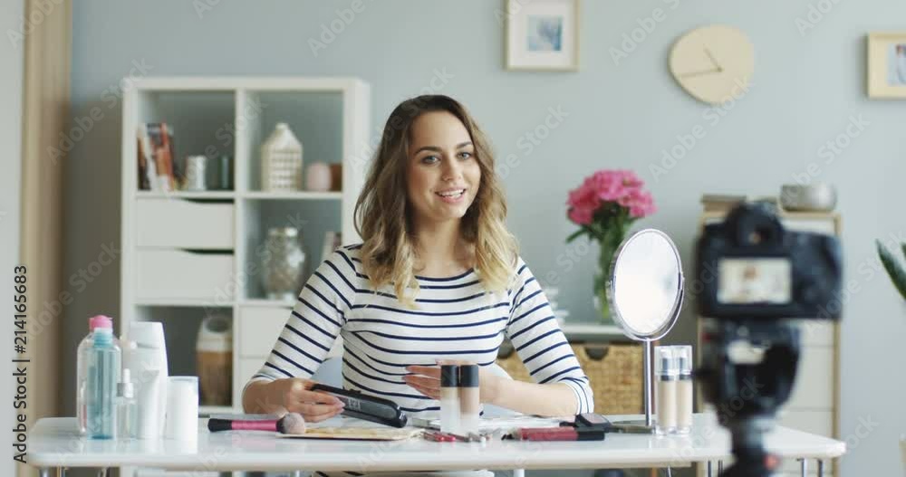 Smiling pretty young woman recording her beauty video blog while talking about choosing eye shadows. Indoors. Smiling pretty young woman recording her beauty video blog while talking about choosing eye shadows. Indoors.