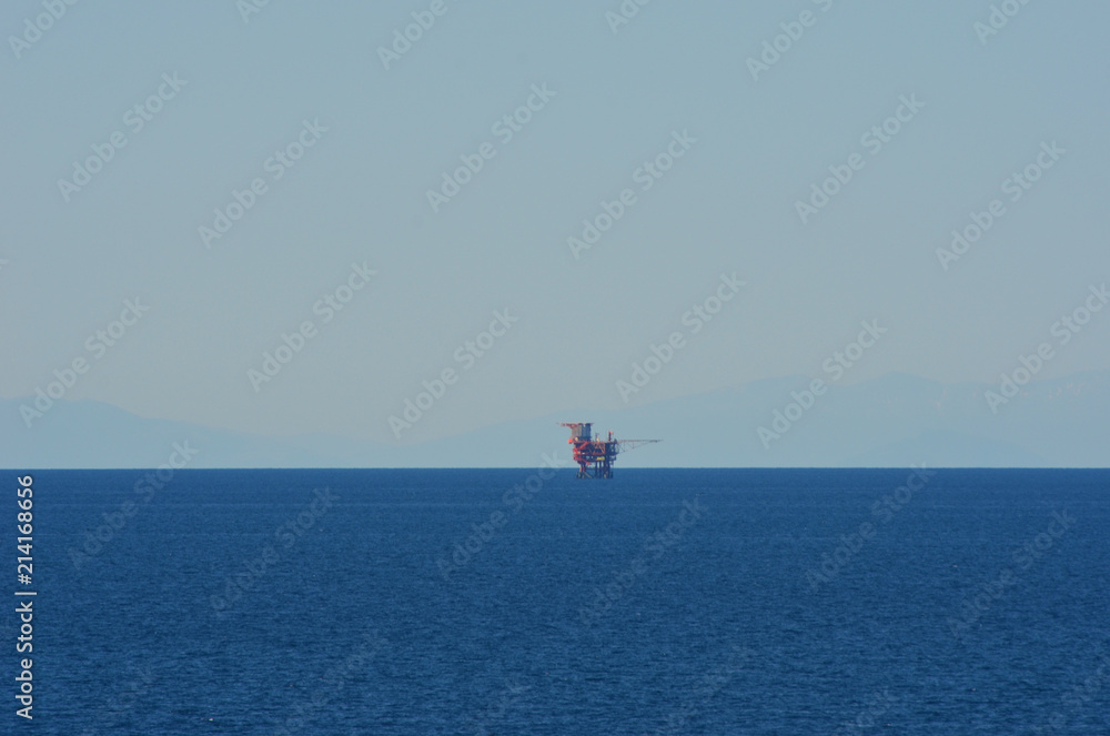 An oil rig, red in colour, is dwarfed by the vastness of the Mediterranean Sea. The sky is blue, with the faint outline of hills in the distance. An oil rig, red in colour, is dwarfed by the vastness of the Mediterranean Sea. The sky is blue, with the faint outline of hills in the distance.