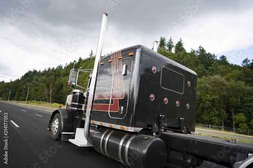 Black classic big rig semi truck with vertical pipes going on wide multiline highway