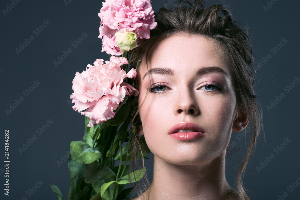 close-up portrait of beautiful young woman with pink eustoma flowers behind ear looking at camera isolated on grey