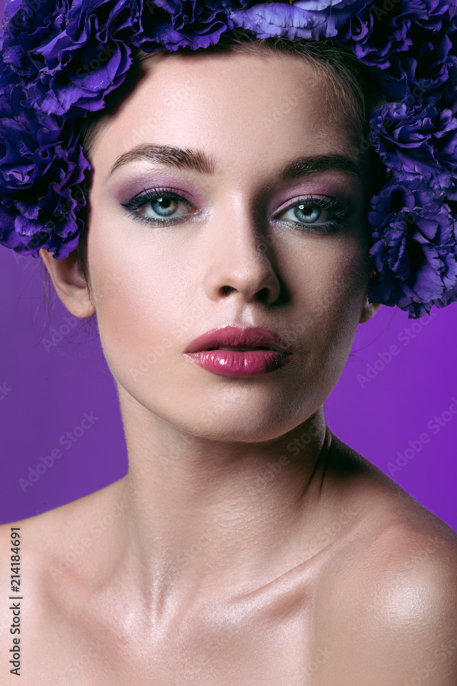 close-up portrait of beautiful young woman with eustoma flowers wreath on head looking at camera isolated on purple
