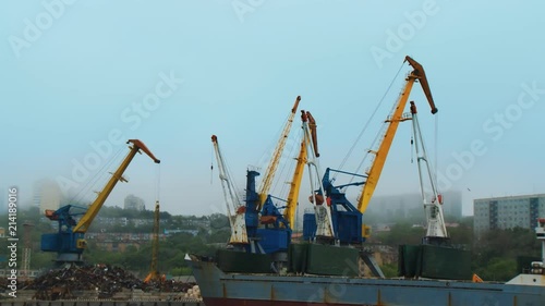 In the seaport of the city of Vladivostok, the crane is unloading scrap-iron from the barge at the pier. The mist closed the part of the city