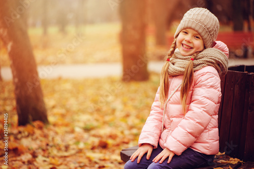 autumn portrait of smiling child girl sitting on bench in park in warm knitted hat and scarf  enjoying outdoor walk in sunny day