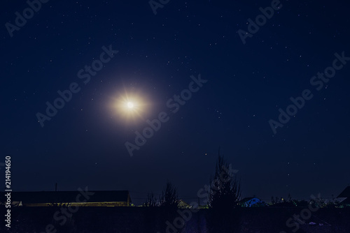The moon and stars in the night sky over the roofs of village houses and trees.