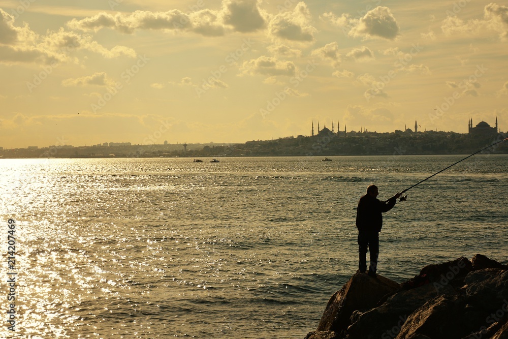 A man is fishing on the rocks by the city silhouette