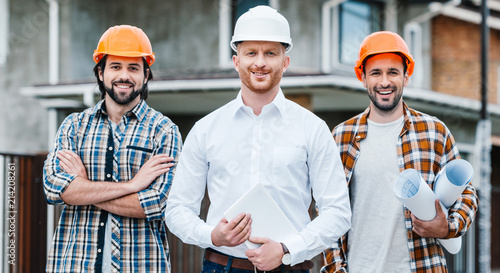 Photo group of successful architects in hard hats looking at camera in front of buildi