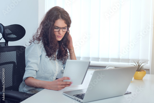 Successful serious business woman sitting at white desk in modern office and reading report document, copy space