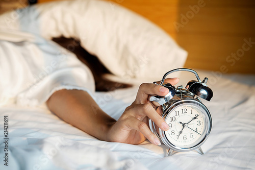 Irritated young woman putting her alarm clock off in the morning with soft morning light. Relaxing concept.