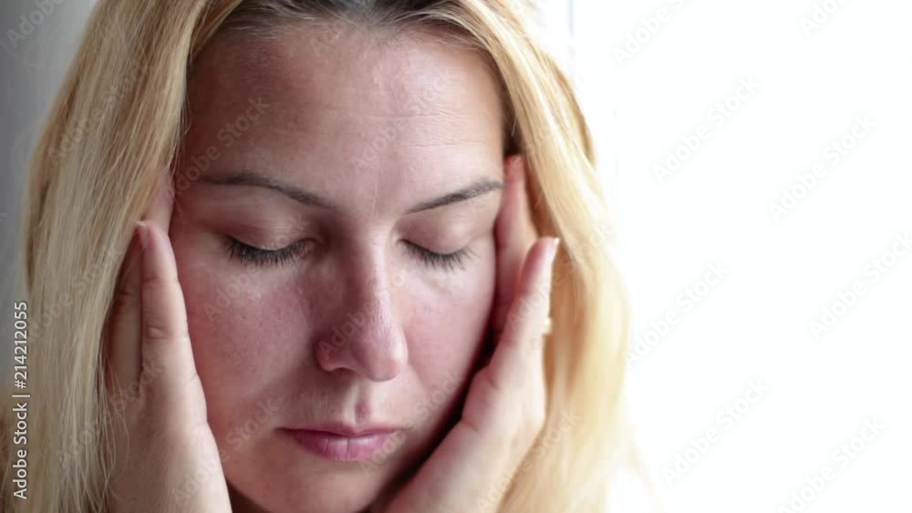 Close up of woman with eyes closed massaging her head because of a headache. Blonde hair sitting at the window Close up of woman with eyes closed massaging her head because of a headache. Blonde hair sitting at the window
