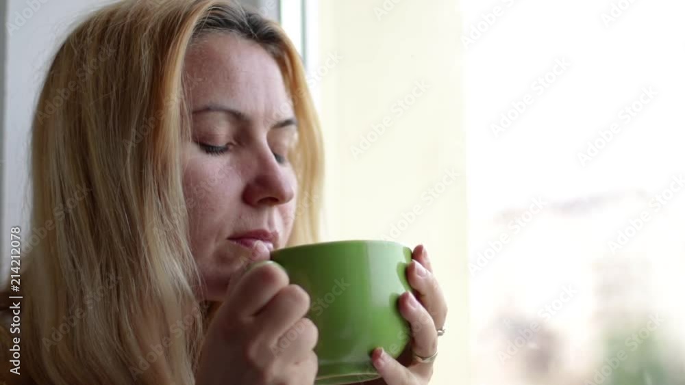 Close up of woman sitting at the window and drinking a hot beverage. Blonde hair lady drinks hot tea Close up of woman sitting at the window and drinking a hot beverage. Blonde hair lady drinks hot tea