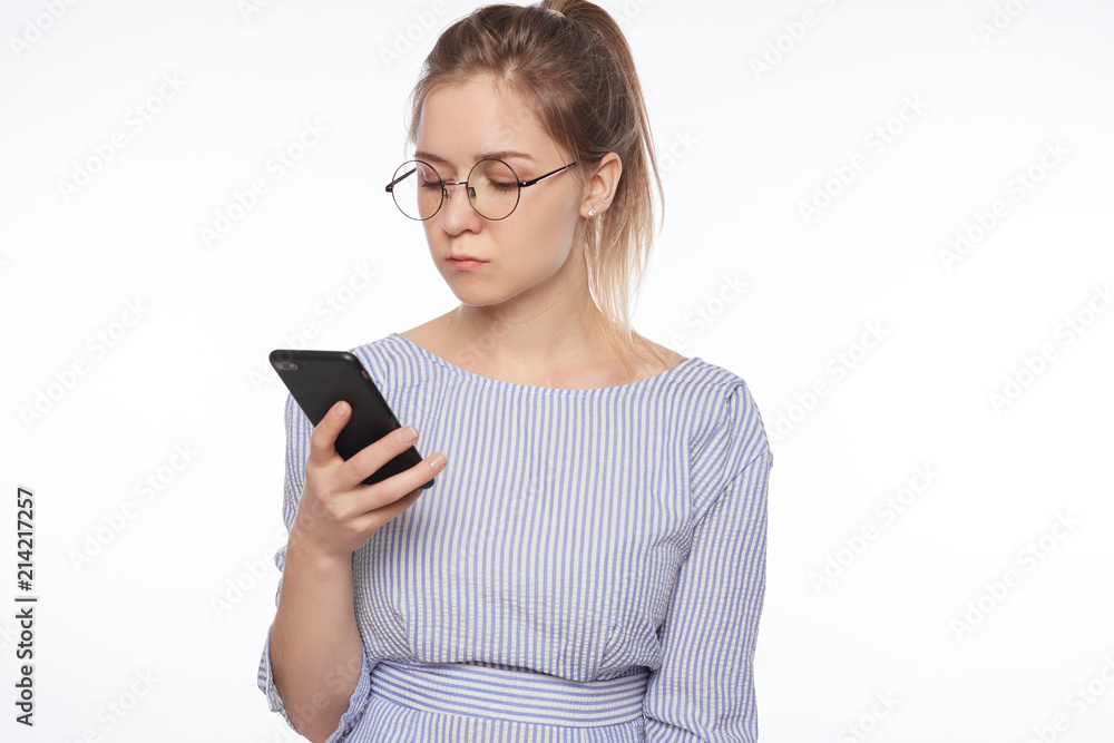 Portrait of attractive young Caucasian woman in spectacles using high-speed Internet connection, texting friends via social networks on smart phone, standing in stripped shirt over white studio wall. Portrait of attractive young Caucasian woman in spectacles using high-speed Internet connection, texting friends via social networks on smart phone, standing in stripped shirt over white studio wall.