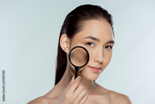 beautiful asian girl with pure skin holding magnifier, skin care concept, isolated on grey