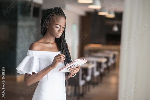 Pretty girl with shiny skin wearing beautiful white dress and standing in a cafe or a restaurant. African american woman with the tablen in her hands. photo