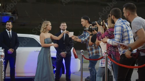 Cheerful blonde woman in gray dress getting out of limousine and giving autographs to fans on red carpet