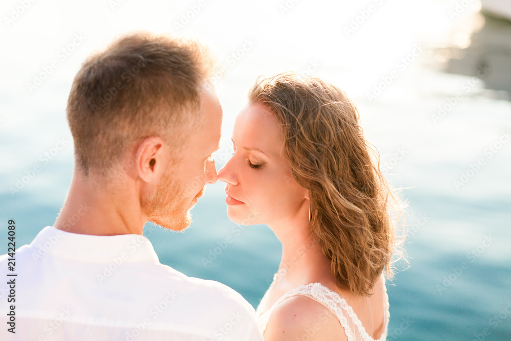 Couple of travelers are sitting by blue sea during romantic honeymoon vacation at sunset after wedding. Man and woman are happy tourists in resort. Lovers are wearing in white clothes outdoor.