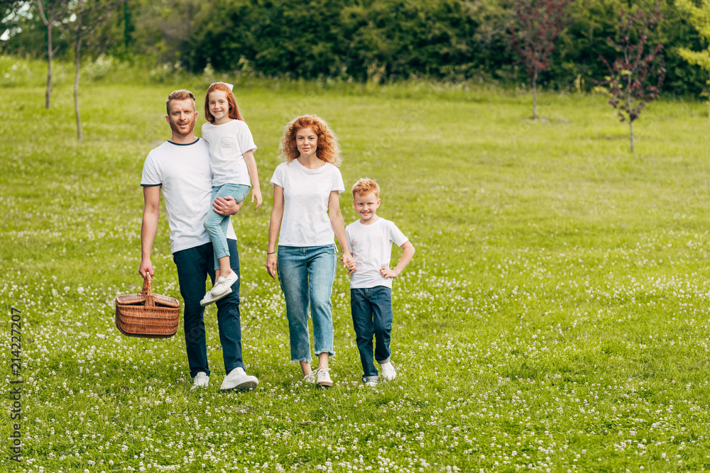 Fototapeta premium happy family smiling at camera while walking with picnic basket in park