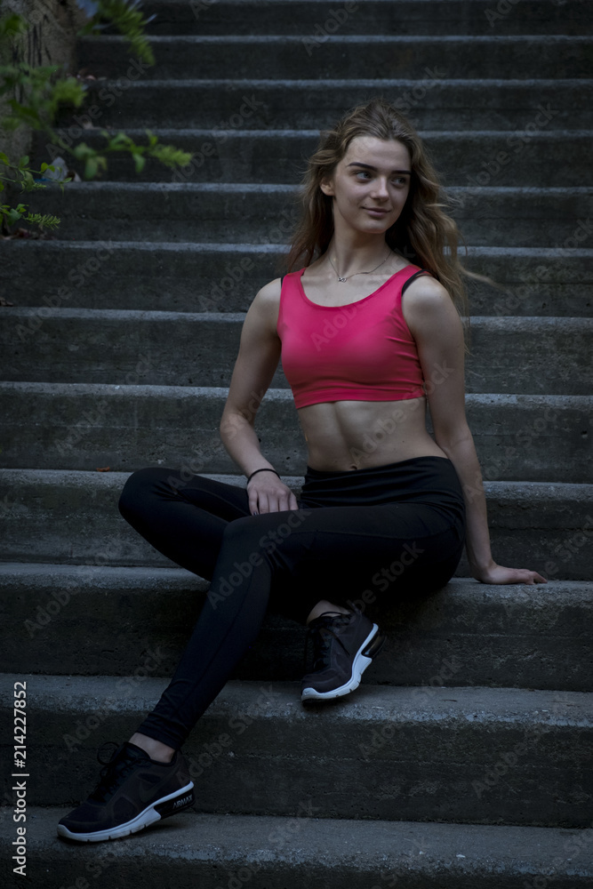 Fototapeta premium Young pretty sportive girl is sitting on the steps on the late summer evening looking down smiling