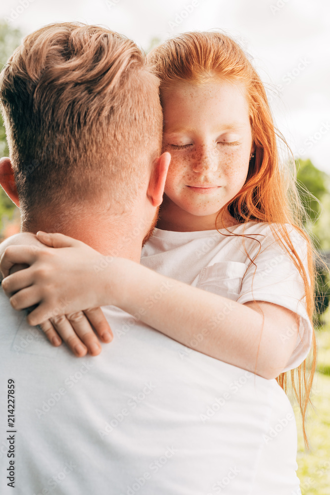 adorable redhead child with closed eyes hugging father at park adorable redhead child with closed eyes hugging father at park
