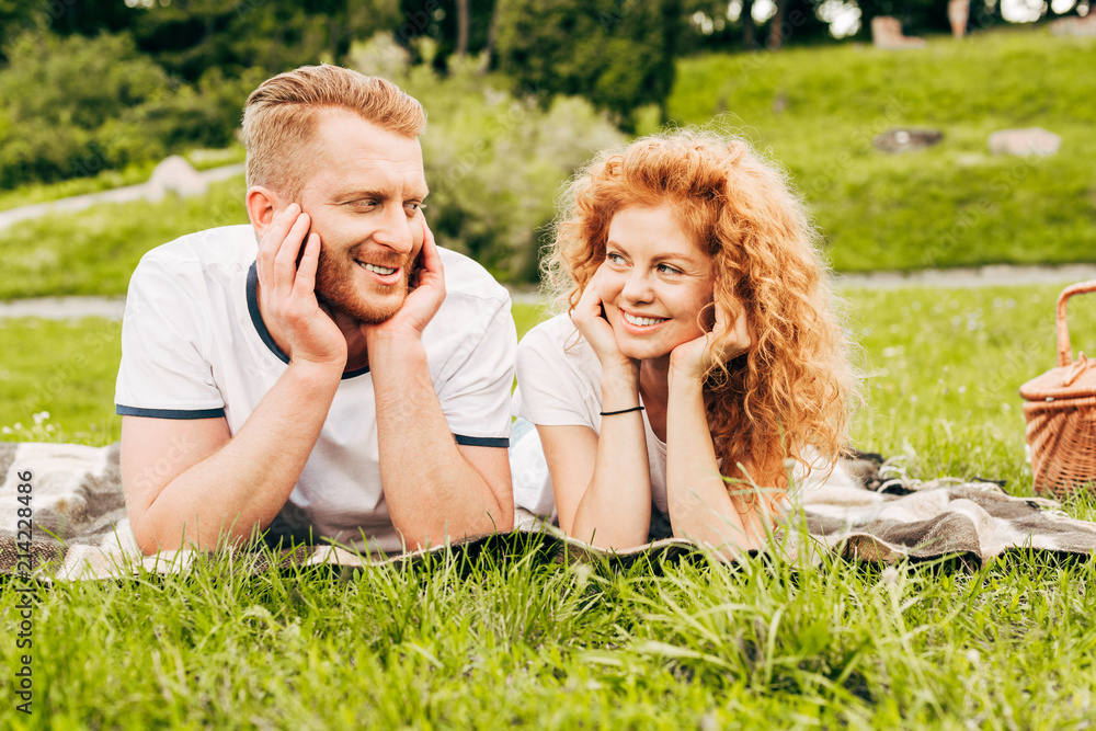 happy redhead couple smiling each other while lying together on plaid at picnic