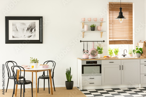 Painting in a black frame in a dining room interior with a table and chairs next to a kitchen interior with tools, plants and cupboards, Real photo