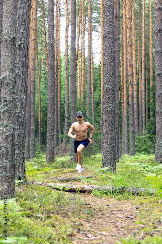 A young man with a muscular bare chest, in shorts and a cap, runs along a pine forest against the background of tree trunks © Makeev Petr