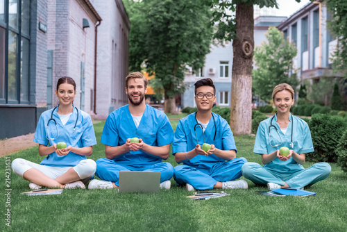 happy multicultural medical students sitting on grass and holding apples