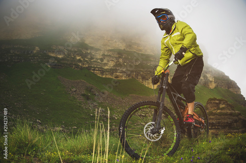 An adult mtb cyclist on a mountain bike at the foot of a cliff surrounded by green grass. Low clouds. North Caucasus. Russia