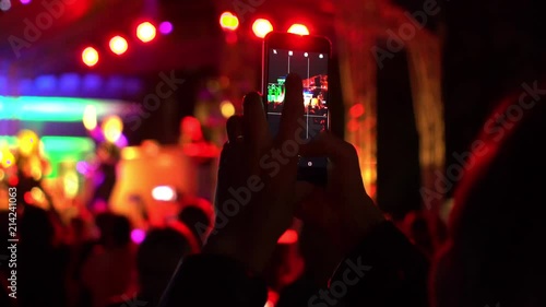 Girl capturing concert on smartphone, large group of happy people enjoying rock concert, clapping with raised up hands, Original multi colored lights blurred background from stage